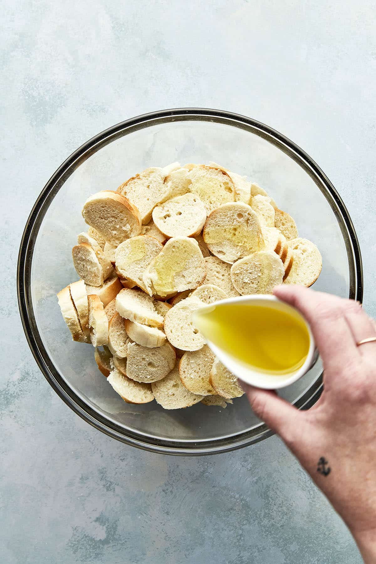 A hand holding a small measuring cup and pouring oil over a bowl of sliced bagel pieces.
