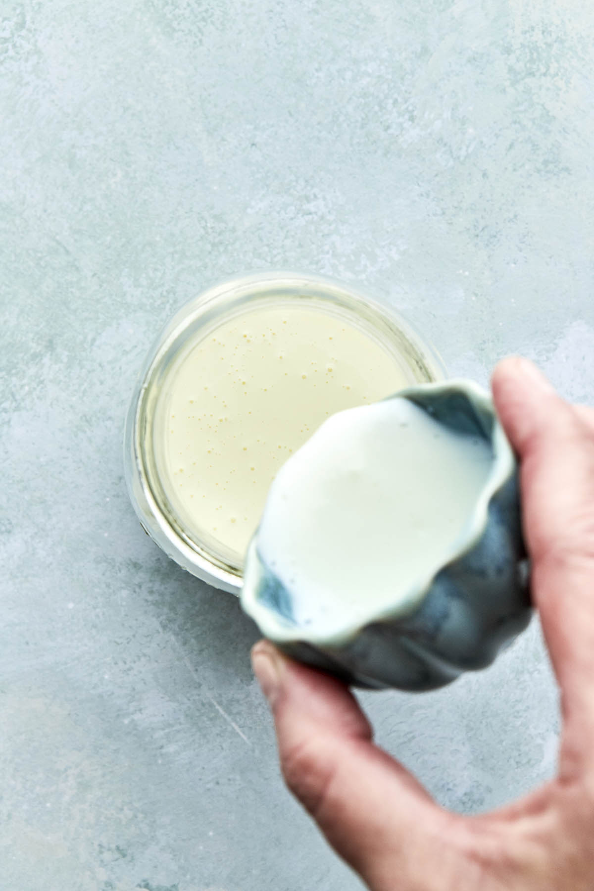 A hand pouring buttermilk from a small blue bowl with scalloped edges into a jar of whipping cream.