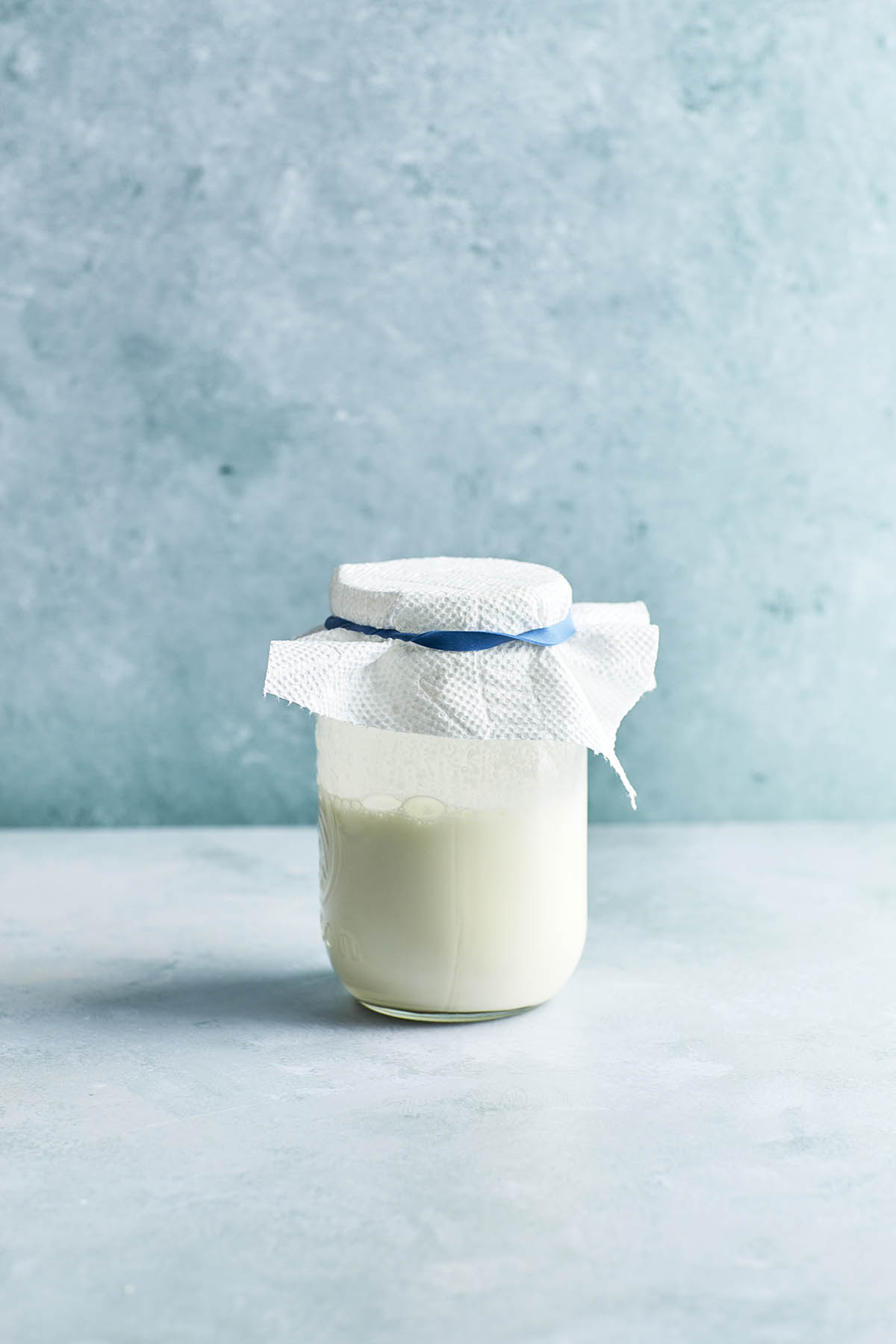 A jar of whipping cream and butter milk shaken together with a paper towel on top secured with a blue rubber band.