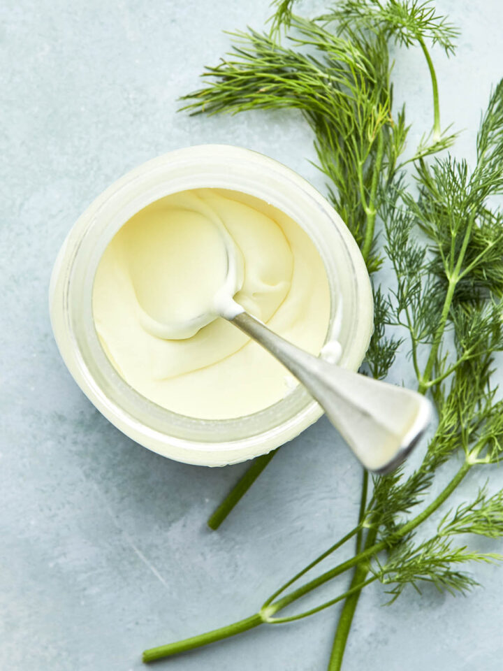 A jar of crème fraîche with a spoon inside the jar and fresh dill laying on the surface next to the jar.