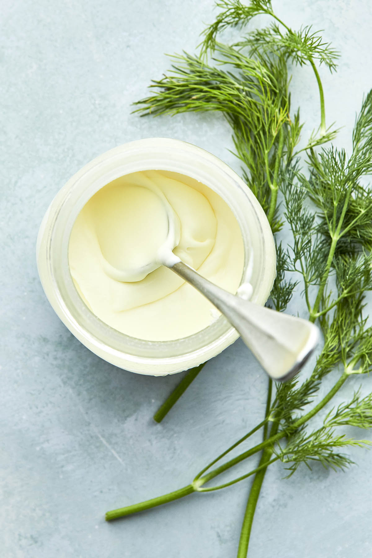 A jar of crème fraîche with a spoon inside the jar and fresh dill laying on the surface next to the jar.
