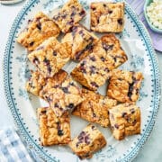 Blueberry blondies on a white and blue oval platter with white chocolate chips, frozen blueberries, plates, and a blue and white checked napkin nearby.