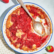 A strawberry cobbler cake in a white rounf baking dish with blue trim, a serving missing and spoon in the dish, and strawberries on the counter nearbly.