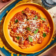 A bowl of lasagna stew topped with fresh basil on a decorative tray with a glass of water, and a bowl of grated Parmesan nearby.