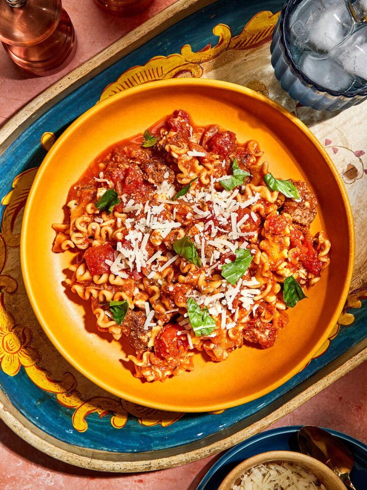 A bowl of lasagna stew topped with fresh basil on a decorative tray with a glass of water, and a bowl of grated Parmesan nearby.