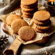Two stacks of chewy molasses cookies, with more cookies laid out on front of the stacks, on a wooden coard surrounded by candles, pine cones, and evergreen.