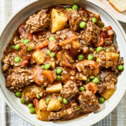 A bowl of hamburger stew on a striped tablecloth with a piece of bread on a plate nearby.