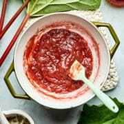 A pot of maple rhubarb jam with fresh rhubarb, cardamom pods, and a small jar of maple syrup nearby.
