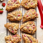 A cooling rack topped with eight strawberry rhubarb cream scones with fresh strawberries and rhubarb stalks nearby.