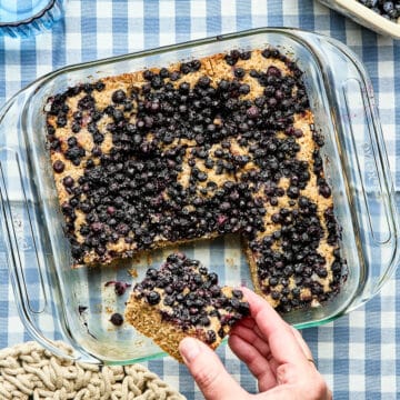 A hand holding a slice of blueberry breakfast cake near the pan of remaining slices on a blue and white checked tablecloth.