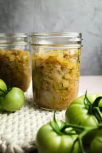 Two jars of green tomato chow with fresh green tomatoes nearby.