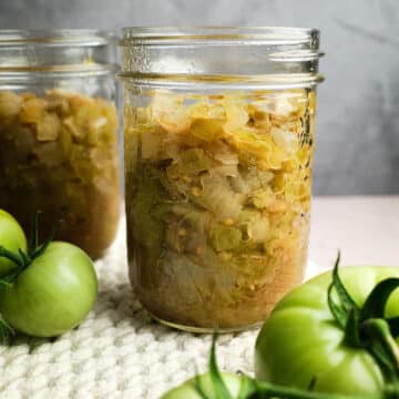 Two jars of green tomato chow with fresh green tomatoes nearby.