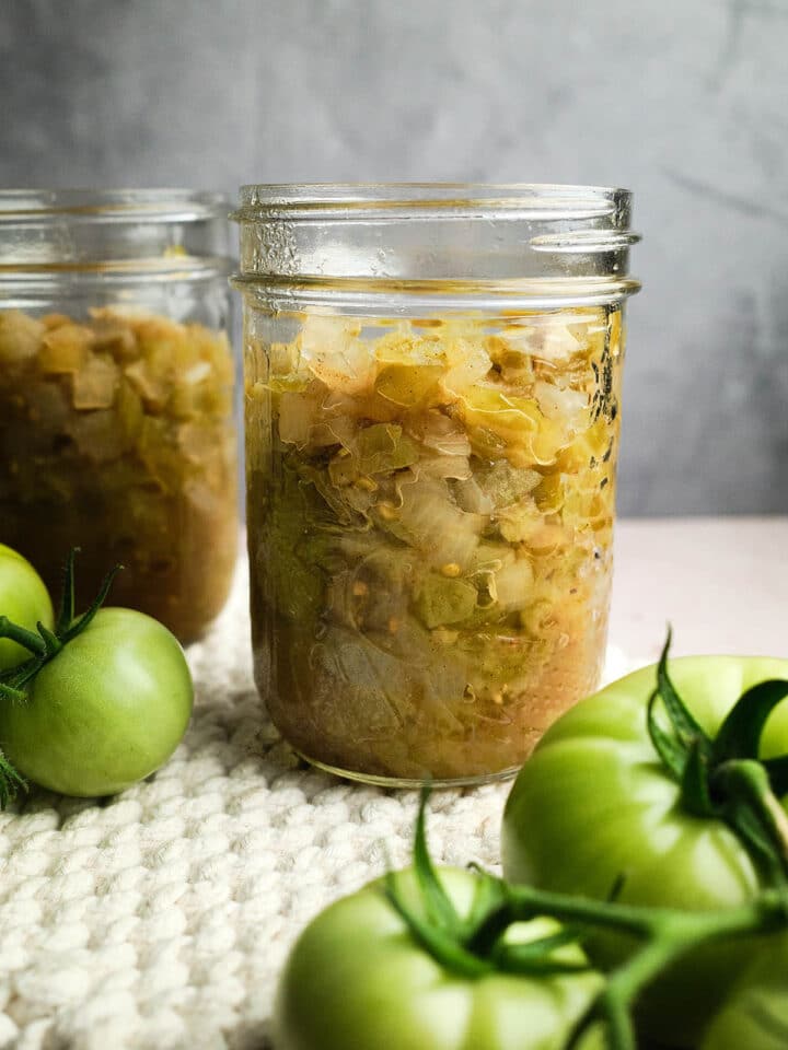 Two jars of green tomato chow with fresh green tomatoes nearby.
