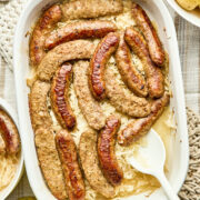 A baking dish of cooked sausage and sauerkraut with a bowl of boiled potatoes, and a jar of mustard pickles nearby.