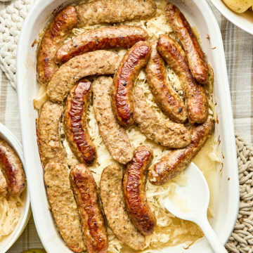 A baking dish of cooked sausage and sauerkraut with a bowl of boiled potatoes, and a jar of mustard pickles nearby.