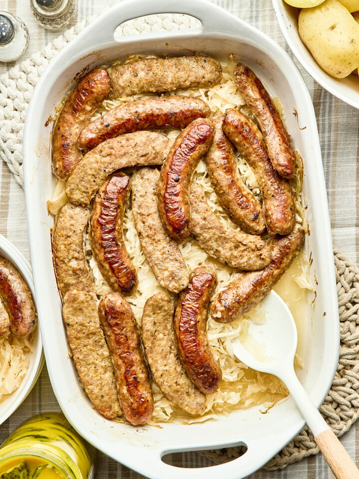 A baking dish of cooked sausage and sauerkraut with a bowl of boiled potatoes, and a jar of mustard pickles nearby.