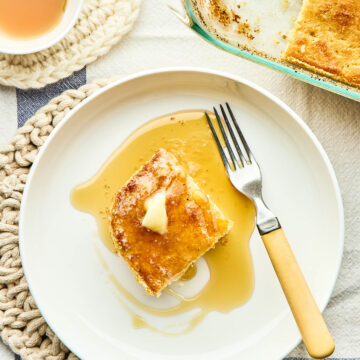 A slice of Johnny cake in a bowl with a yellow-handled fork topped with melting butter and maple syrup with the pan of cake and a small dish of maple syrup nearby.