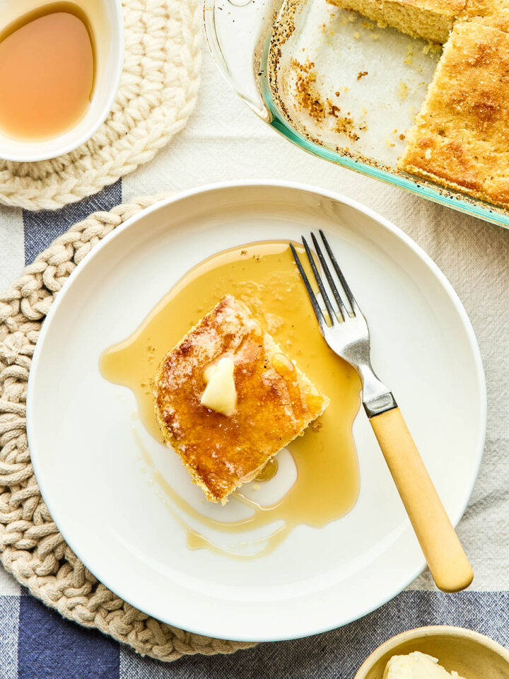 A slice of Johnny cake in a bowl with a yellow-handled fork topped with melting butter and maple syrup with the pan of cake and a small dish of maple syrup nearby.