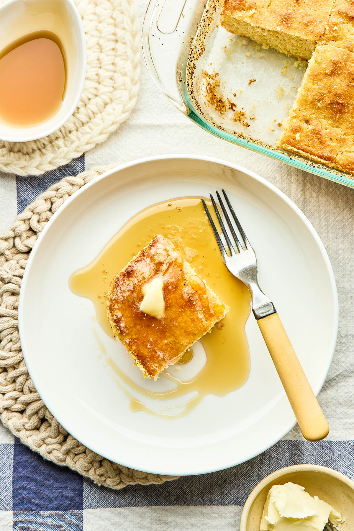 A slice of Johnny cake in a bowl with a yellow-handled fork topped with melting butter and maple syrup with the pan of cake and a small dish of maple syrup nearby.