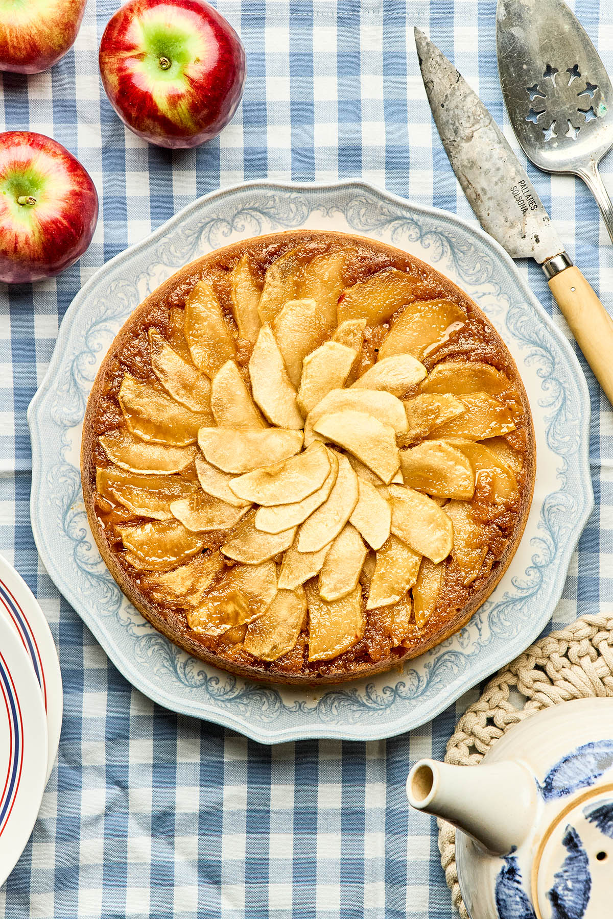 An unsliced apple upside-down cake with maple syrup on a blue and white serving plate on a blue and white checked tablecloth.