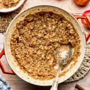 Dutch oven apple crisp on a woven trivet set on a tabletop with a scoop removed to a nearby bowl topped with ice cream.