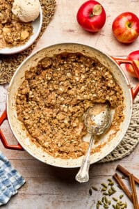 Dutch oven apple crisp on a woven trivet set on a tabletop with a scoop removed to a nearby bowl topped with ice cream.