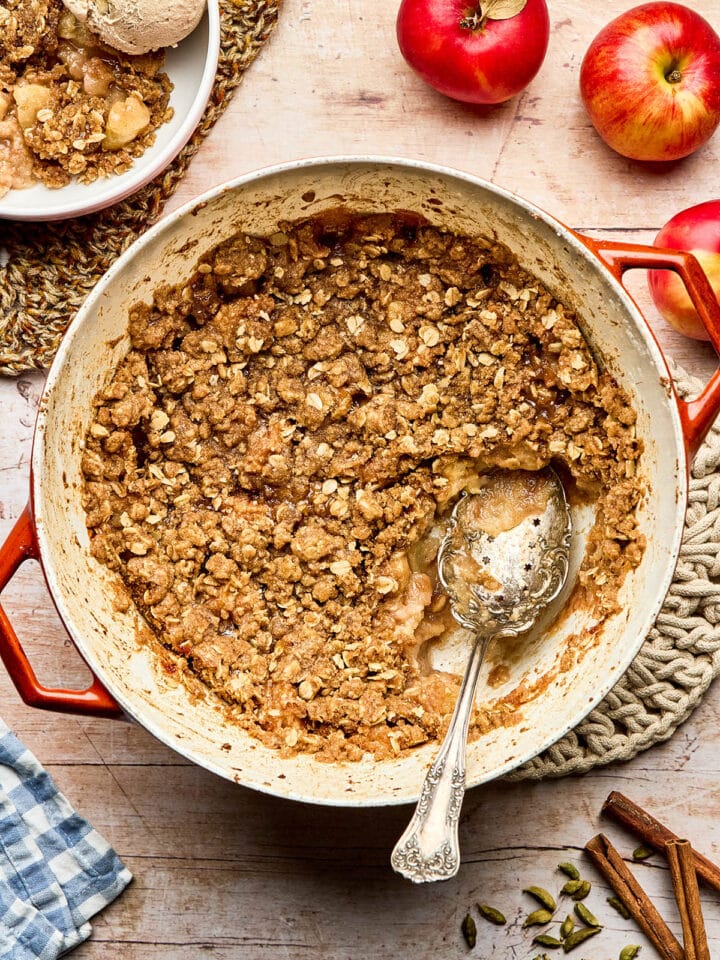 Dutch oven apple crisp on a woven trivet set on a tabletop with a scoop removed to a nearby bowl topped with ice cream.