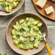 A beige bowl of creamy pesto gnocchi topped with basil and toasted pine nuts, set on a textured linen cloth with a yellow-handled fork and shaved Parmesan on a wooden board.