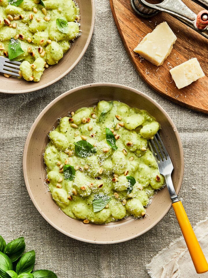 A beige bowl of creamy pesto gnocchi topped with basil and toasted pine nuts, set on a textured linen cloth with a yellow-handled fork and shaved Parmesan on a wooden board.