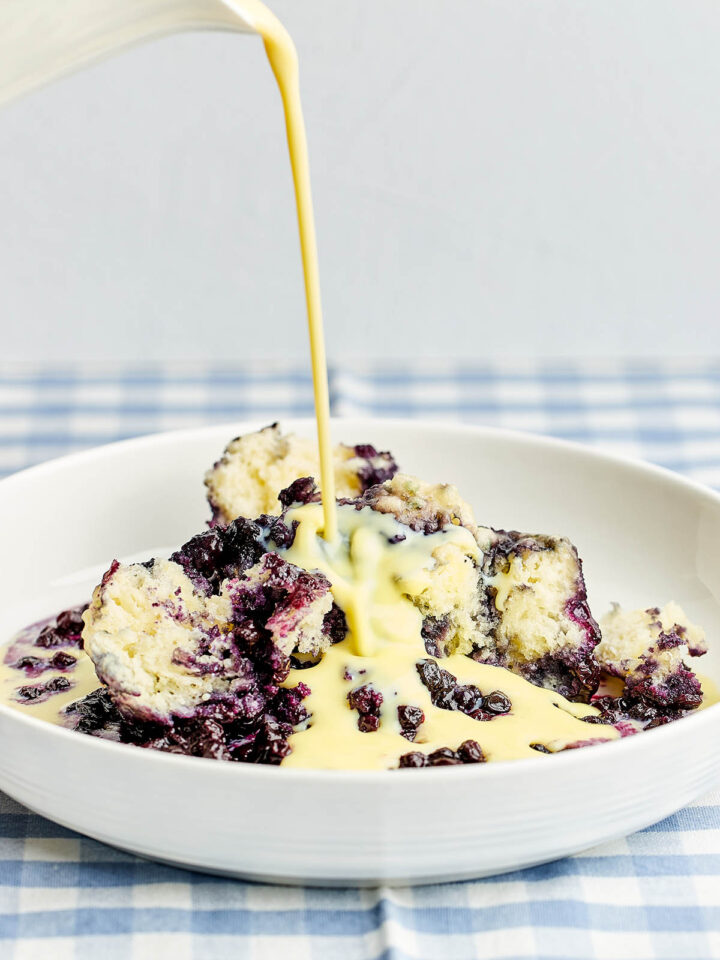 Vanilla custard sauce being poured over a bowl of Nova Scotia blueberry grunt.