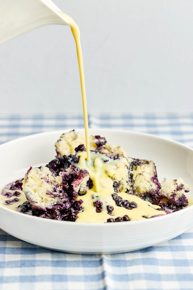 Vanilla custard sauce being poured over a bowl of Nova Scotia blueberry grunt.