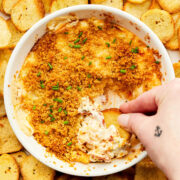 A hand dipping a bagel chip into a dish of hot lobster dip with cream cheese.
