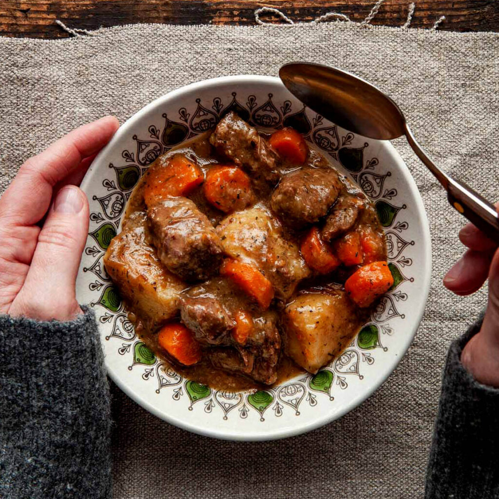 A woman's hands holding a spoon about to dig into a bowl of Dutch oven beef stew.