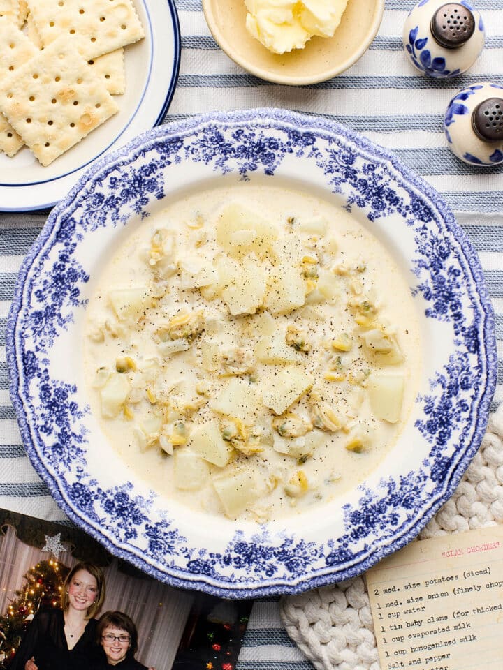 A bowl of my Mum's clam chowder (using canned clams) on a table with saltine crackers, butter, a photo of me and my Mum, and her original recipe card from the 1970s nearby.